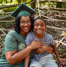 Commencement_Skylar-Tirotta Smiling woman in graduation cap embraces child while sitting on a netted playground structure in a wooded area.