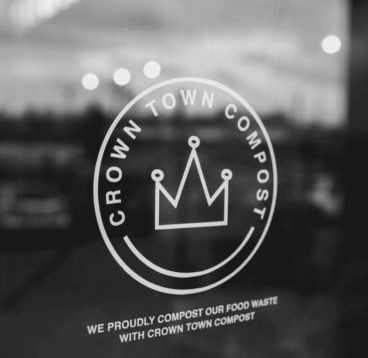 Crown Town Business logo Black and white logo for Crown Town Compost on a glass surface, featuring a stylized crown inside a circle and a caption about composting food waste.