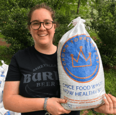 Crown Town Customer A woman smiles while holding a white Crown Town Compost bag with a blue and orange logo and the text “Once food waste, now healthy soil.” She stands outdoors with other compost bags behind her.