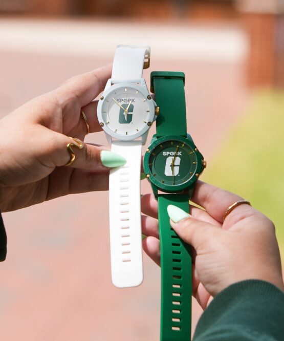 SPGBK-Watch-2 Hands holding two wristwatches — one white and one green — each featuring the UNC Charlotte “All-in-C” logo on the face.