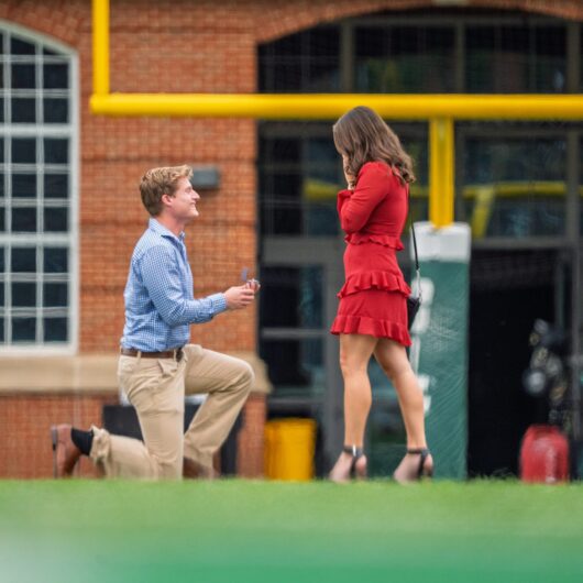 Katerina-Lantz-Niner-Love-Story A man kneels on a football field proposing with a ring to a woman in a red dress, with stadium goalposts and a brick building in the background.