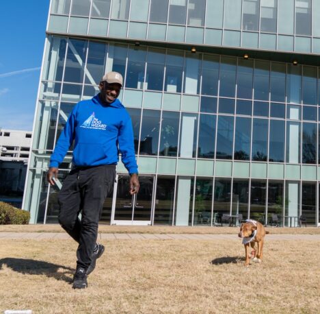 Man in blue hoodie leading the way for his brown dog. With a city high rise in the background.