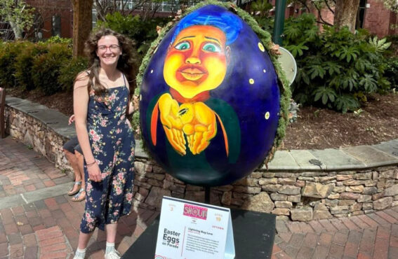 Easter Egg_ Lightning Bug Love. copy Smiling woman in a floral jumpsuit stands beside a large painted Easter egg sculpture during the Easter Eggs on Parade public art exhibit.