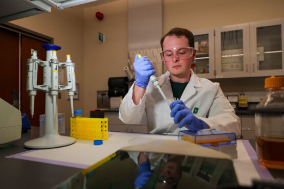 Equity_Brendon_Coats-2 Student in a lab coat and safety goggles uses a pipette to transfer liquid in a laboratory, surrounded by scientific equipment and sample containers.