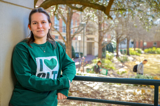 Equity_Lindsay_Rammalaere Student in a green “I Heart CLT” sweatshirt leans on a railing outdoors on campus, with trees, walkways, and other students in the softly blurred background.