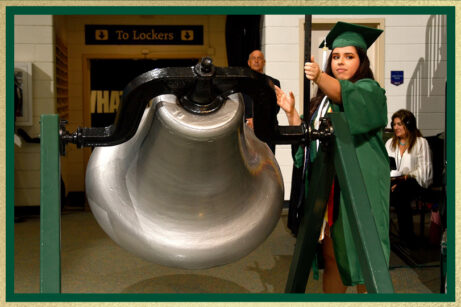Equity_Maria_Garcia-4 Graduate in a green cap and gown rings a large ceremonial bell indoors during a commencement event, with people and signage visible in the background.