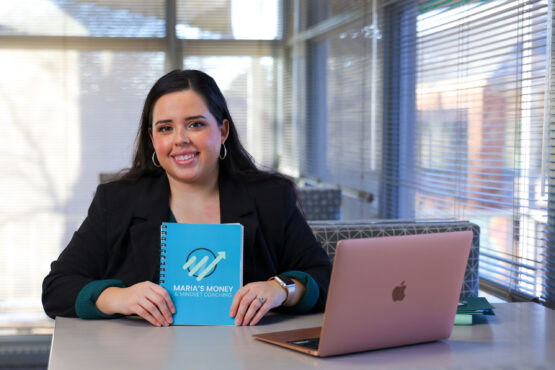 Equity_Maria_Garcia Woman seated at a table in a bright office setting holds a notebook labeled “Maria’s Money & Mindset Coaching,” with a laptop open beside her and window blinds in the background.