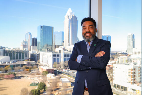 Equity_Micah_Gaddy-2 Professional stands with arms crossed in front of a large window overlooking the Charlotte skyline, with city buildings visible in daylight.