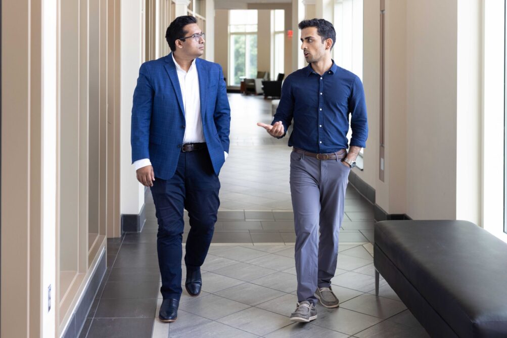 Two men walk and talk in a sunlit hallway.