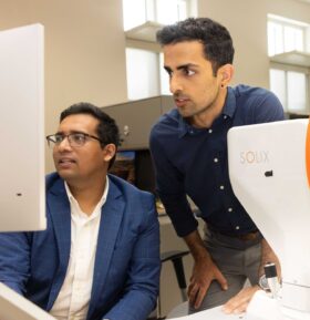 Three people use a eye imaging device in a lab, with one person seated at a computer and another guiding a patient.