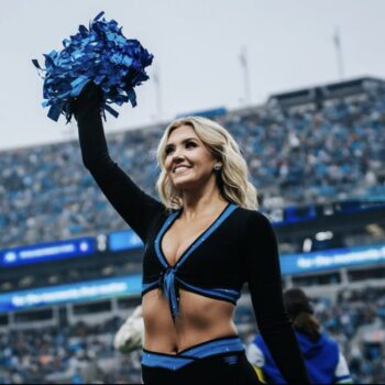 Haygan Hayes, TopCat A cheerleader in a black and blue outfit smiles while holding blue pom-poms overhead on a football field filled with fans.
