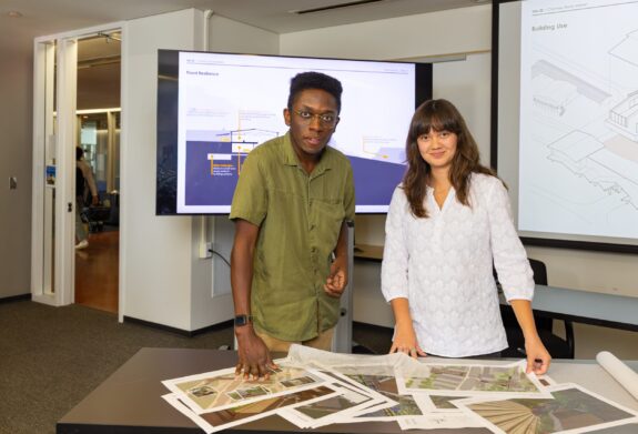 Two students stand at a table with printed design plans, presenting their project in front of screens displaying diagrams and renderings.