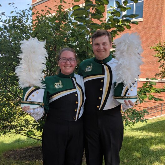Hayley-Owen-Niner-Love-Story Two marching band members in green, black, and gold Charlotte uniforms stand outdoors holding white plumed hats, smiling in front of a campus building.