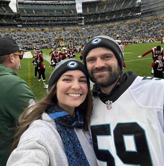 Lauren-Smith-Niner-Love-Story A couple wearing winter hats takes a selfie on the field at Lambeau Field, with a scoreboard and marching band visible in the stadium behind them.