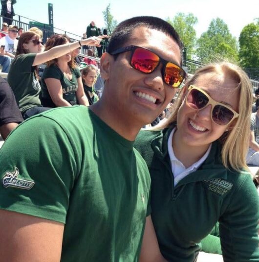 Kate-Daluyin-Niner-Love-Story Two fans wearing green Charlotte apparel smile for a photo in the stands at a sunny outdoor sporting event.