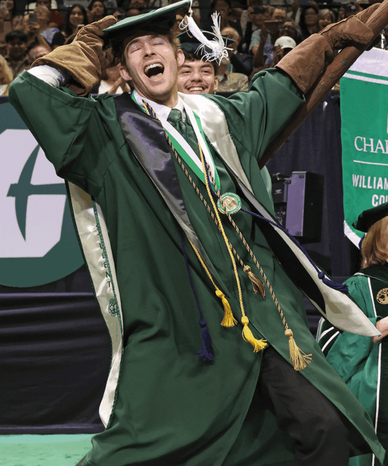 Excited graduate in green regalia and honor cords raises a pickaxe while celebrating.