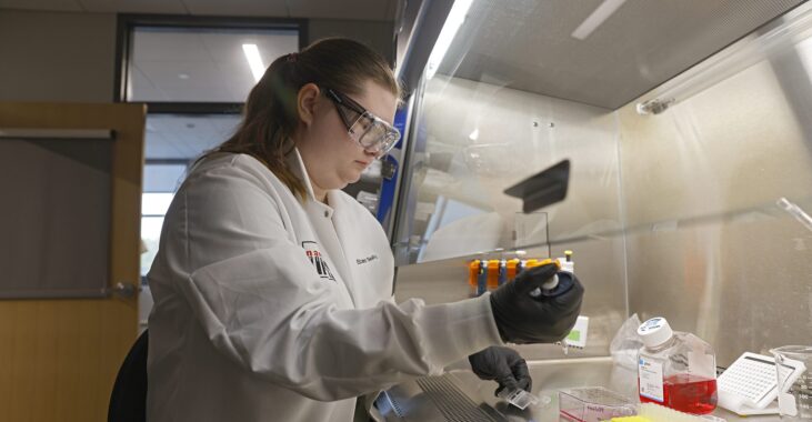 KCOS_Afonin_Lab_24039_6967_kpl Person in a lab coat using a pipette at a lab bench under a fume hood.