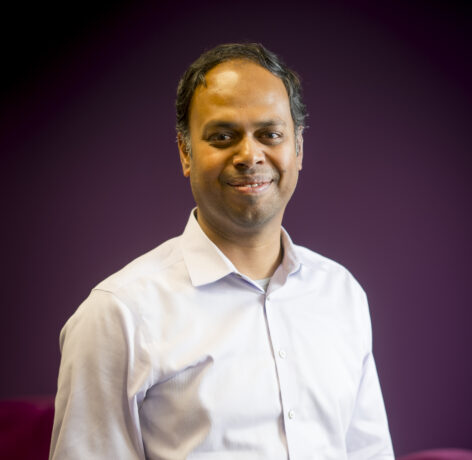 A man wearing a white shirt and blue pants stands confidently in front of a stylish purple backdrop.