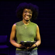 Madison King A UNC Charlotte student smiles on a darkened stage, holding a tablet under theatrical lighting.