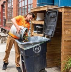 Man-dumping-trash-2 A man in an orange shirt and brown pants pours a bucket of food scraps into a black compost bin labeled with the Crown Town Compost logo, standing beside a wooden storage area outside a brick building.