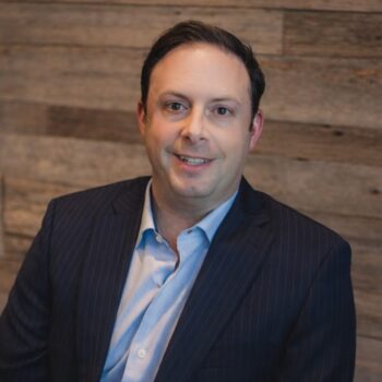 Mark Lambardi Professional headshot of a man in a dark pinstripe suit and light blue shirt, seated in front of a wooden wall background.