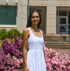 Maya Barbee CHESS May 25-2 A UNC Charlotte student stands in front of pink flowering bushes and a campus building on a sunny day.