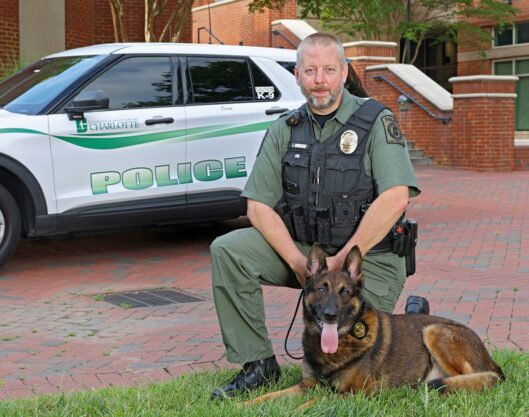 PPS_Group_Photos_25035_5815_kpl A police officer kneels beside a German Shepherd in front of a campus police SUV.