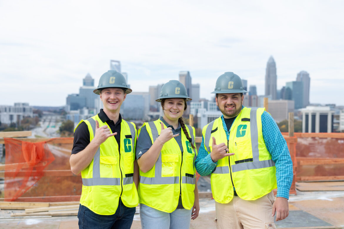 Three construction students stand in hard hats in front of the Charlotte skyline