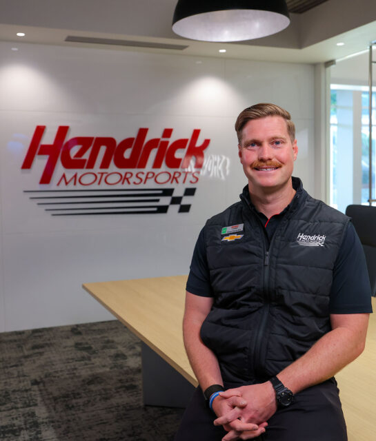 A Hendrick Motorsports performance engineer sits at a conference table in a modern meeting room, smiling in front of a Hendrick Motorsports logo displayed on the wall.