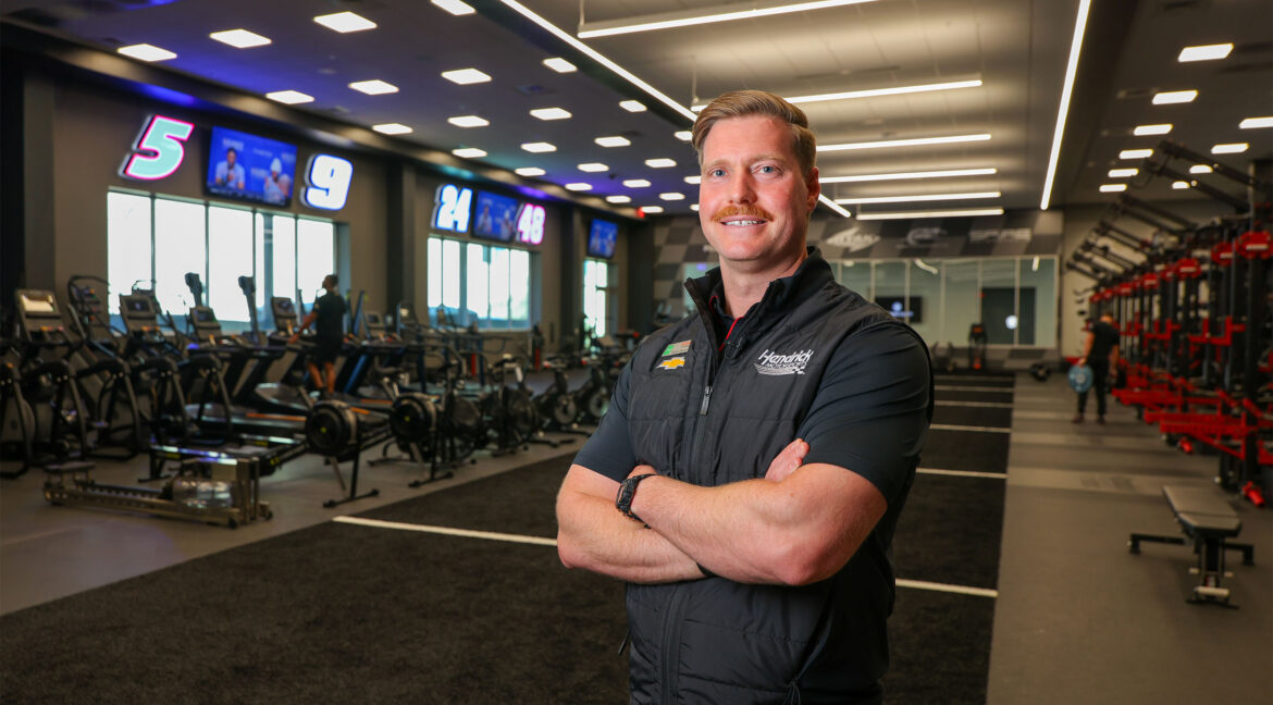 A Hendrick Motorsports staff member stands with arms crossed inside a modern training and performance facility equipped with cardio machines, weightlifting stations, and digital timing displays.
