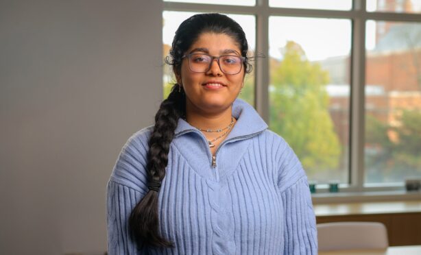 A student wearing glasses and a light blue sweater stands indoors in front of a large window, with campus buildings and trees visible in the background.