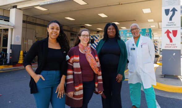 Four people, including a medical professional in a lab coat, stand smiling outside a hospital emergency entrance, with an ambulance visible in the background.