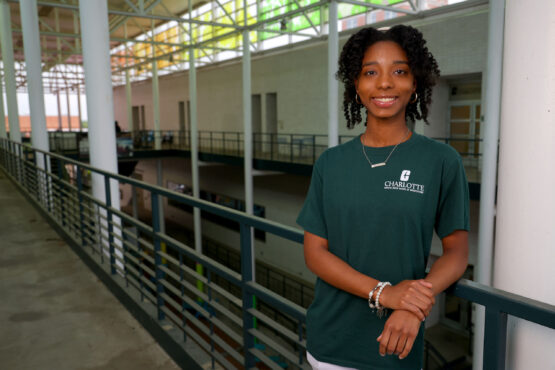 Serenity_Snead_equity-engine Smiling student in a green UNC Charlotte T-shirt stands in a modern campus atrium, leaning on a railing with open walkways and structural beams in the background.