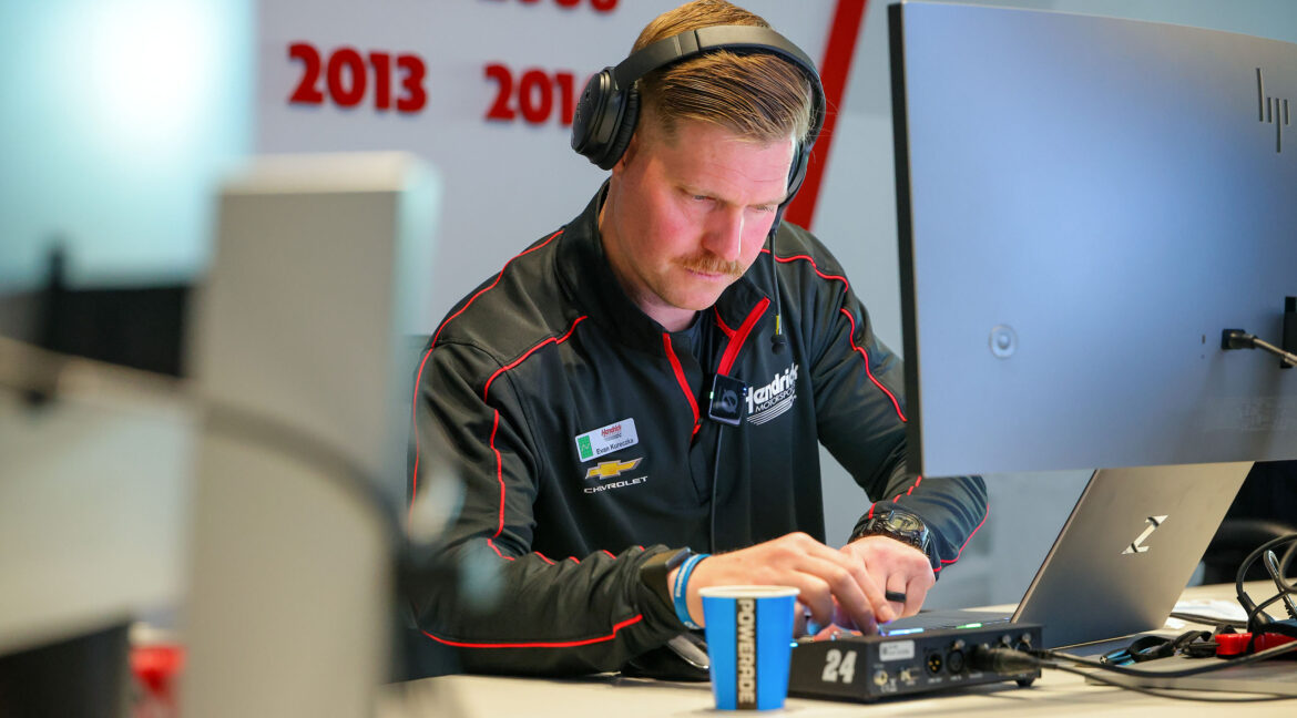 A Hendrick Motorsports performance engineer wearing a headset works at a computer workstation, monitoring data and preparing race operations.