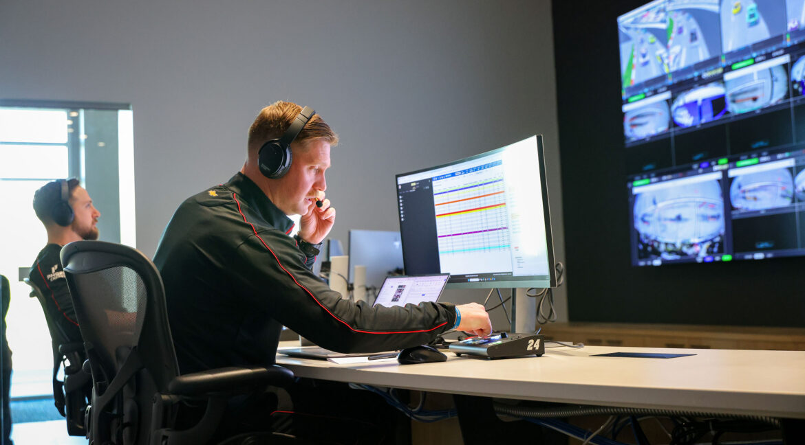 A Hendrick Motorsports engineer uses a headset and multiple monitors in a race operations center while reviewing live timing, strategy data, and track camera feeds.
