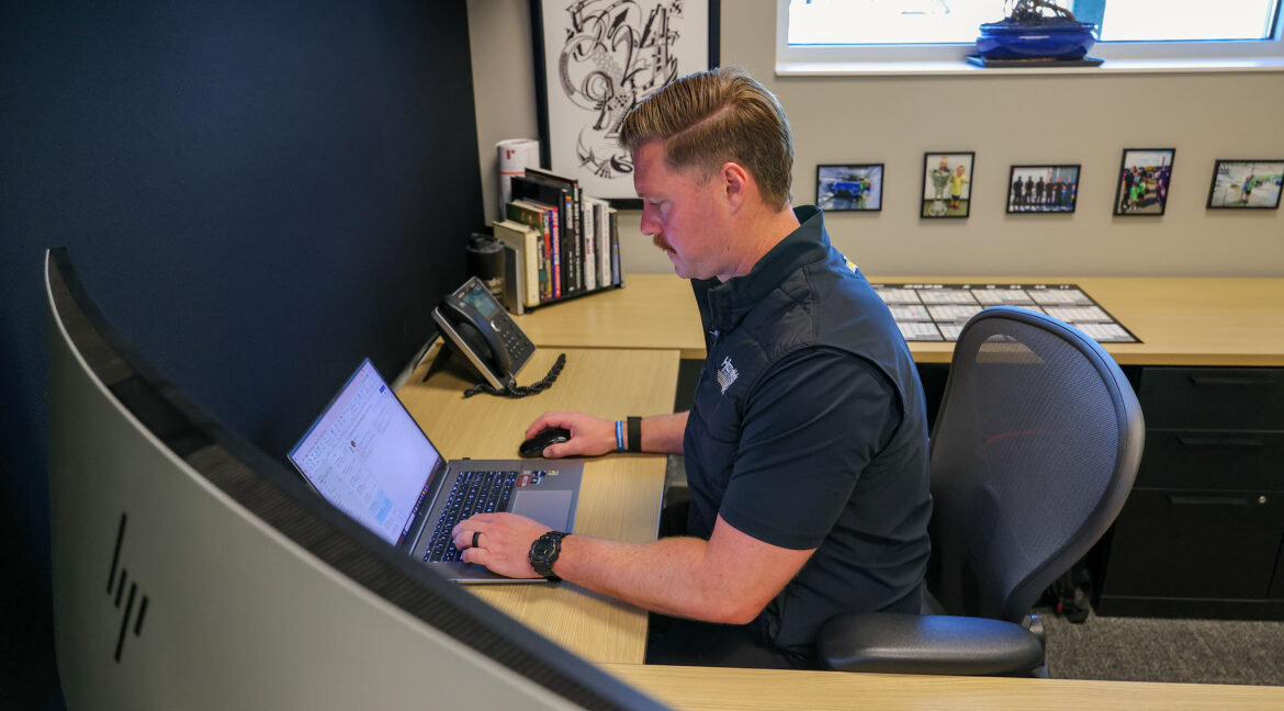 A Hendrick Motorsports engineer works at a laptop in a quiet office, analyzing performance data at a desk with books, framed photos, and a telephone.