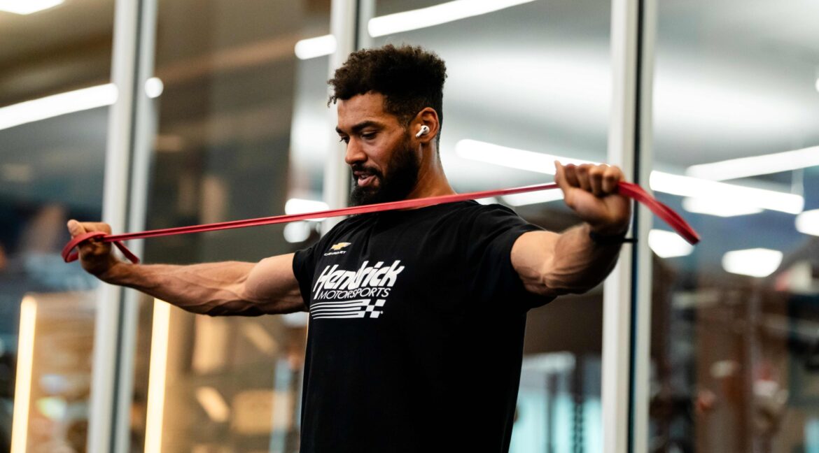 A Hendrick Motorsports athlete performs a resistance band exercise inside a modern training facility to improve upper-body strength and mobility.