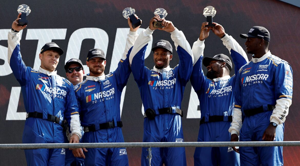 Members of the Hendrick Motorsports team stand on a podium holding trophies after a race, celebrating their achievement together.