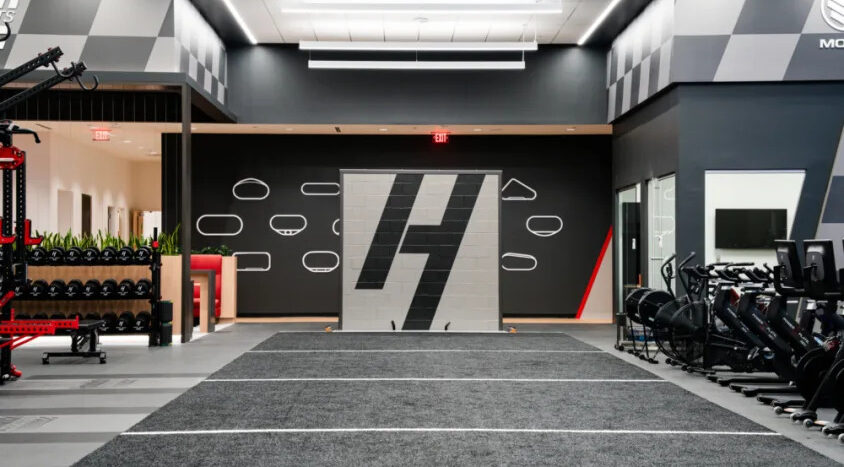 Modern Hendrick Motorsports training facility with indoor turf lanes, strength equipment, cardio machines, and a large Hendrick logo on the far wall.
