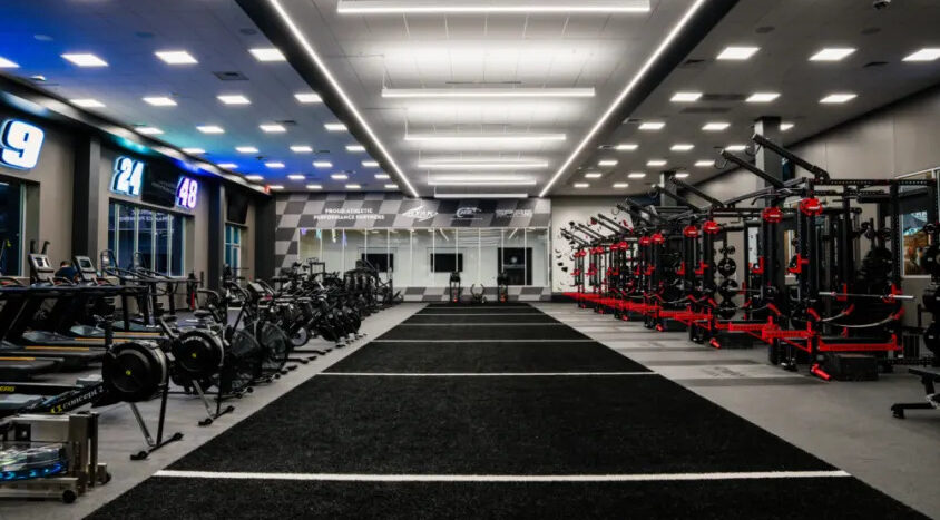 Wide view of the Hendrick Motorsports performance center featuring indoor sprint lanes, weight racks, rowing machines, and cardio equipment in a spacious gym.
