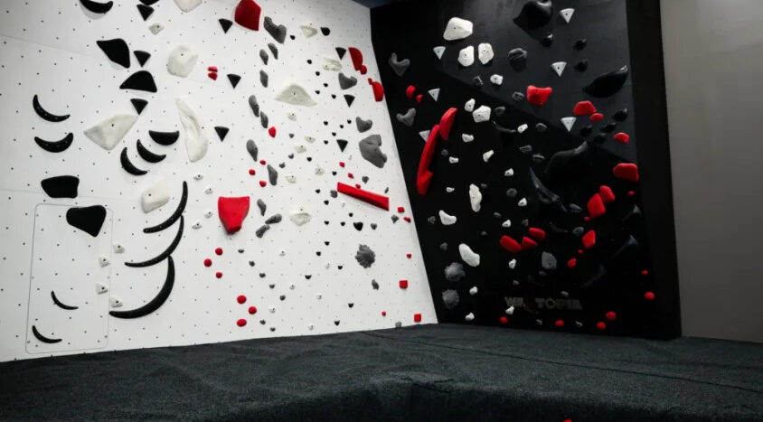 Indoor bouldering wall with black, white, gray, and red climbing holds above padded flooring for strength and agility training.