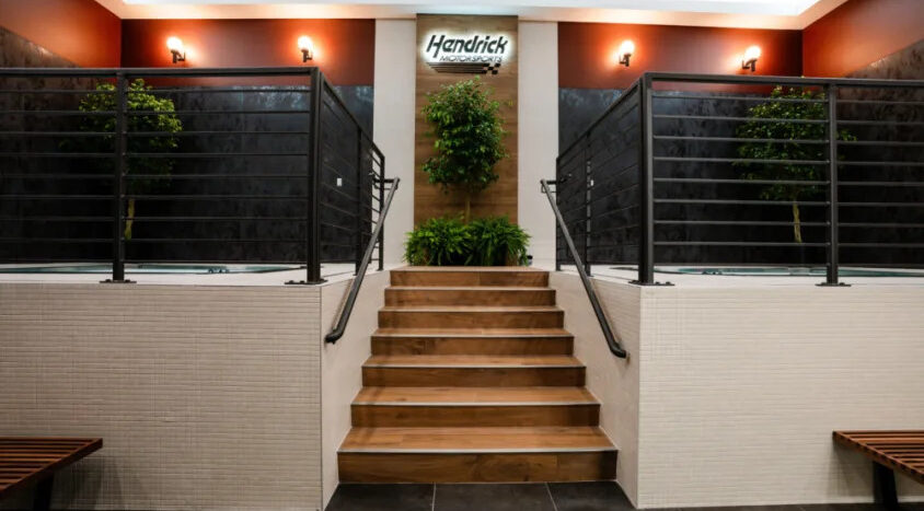 Lobby inside Hendrick Motorsports featuring a central staircase, indoor plants, and modern architectural finishes leading to the main entrance.