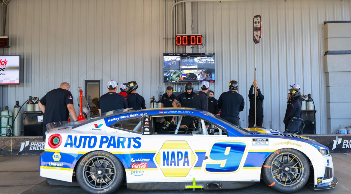 The No. 9 NAPA Auto Parts Chevrolet sits in the garage while the Hendrick Motorsports pit crew prepares for practice or a race under a timing display and monitor wall.
