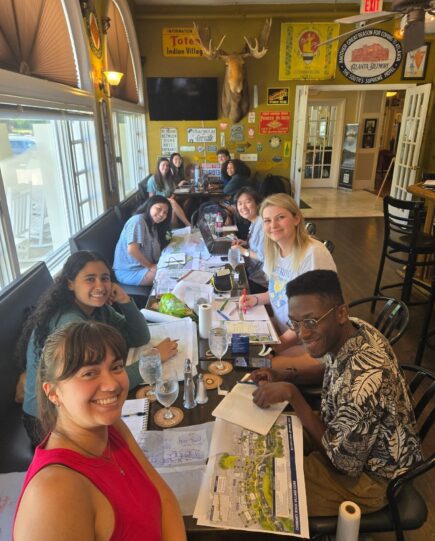 Summer Studio A group of students gathered around a long table in a restaurant covered with sketches, laptops, and maps. They are smiling and working together, with eclectic wall décor and a mounted moose head in the background.