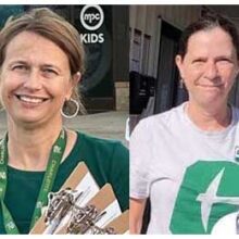 Two women smile while holding clipboards outdoors, both wearing UNC Charlotte gear and lanyards.