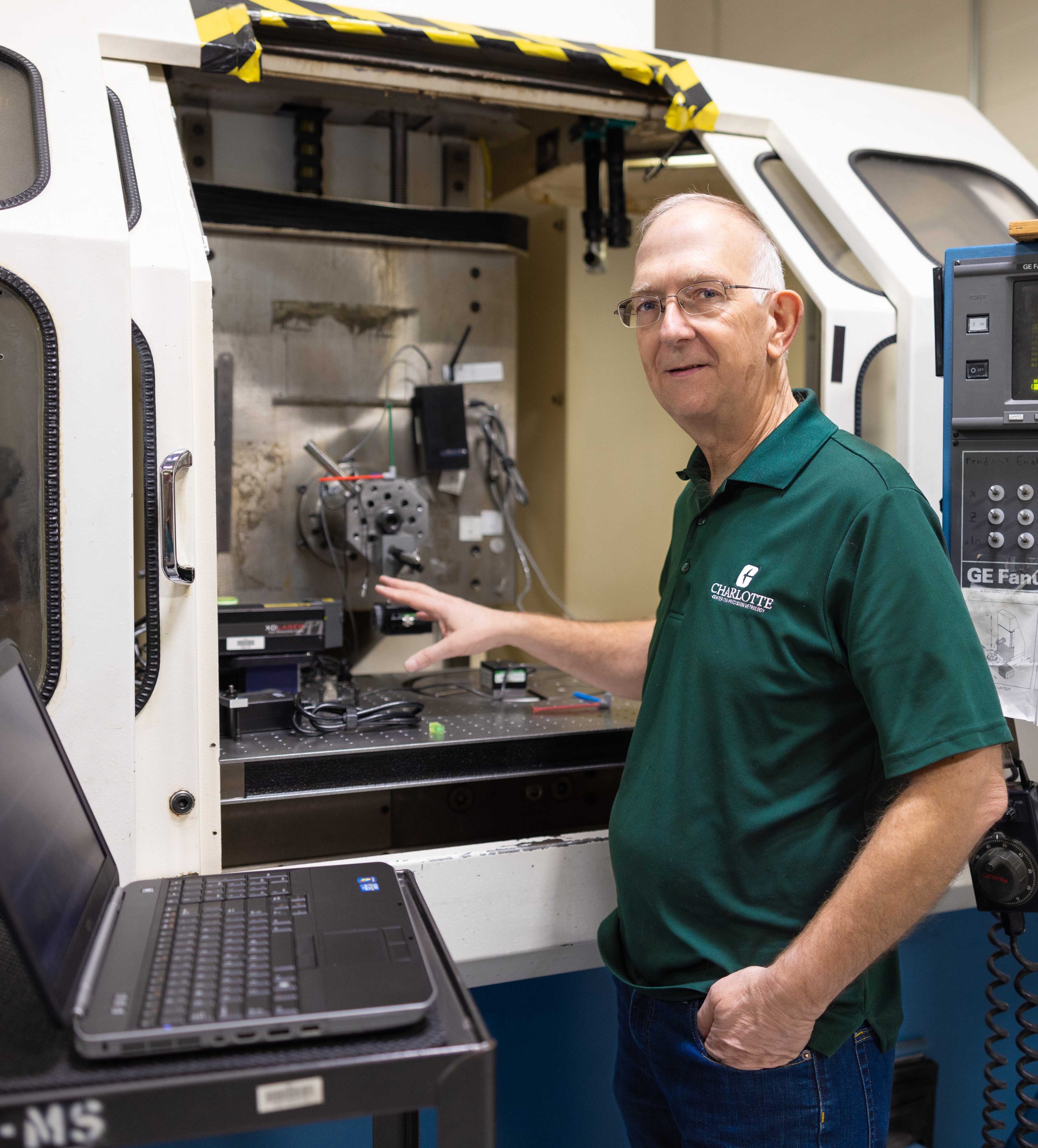 Man in a green polo demonstrates equipment in a lab, standing beside an open machine and laptop.