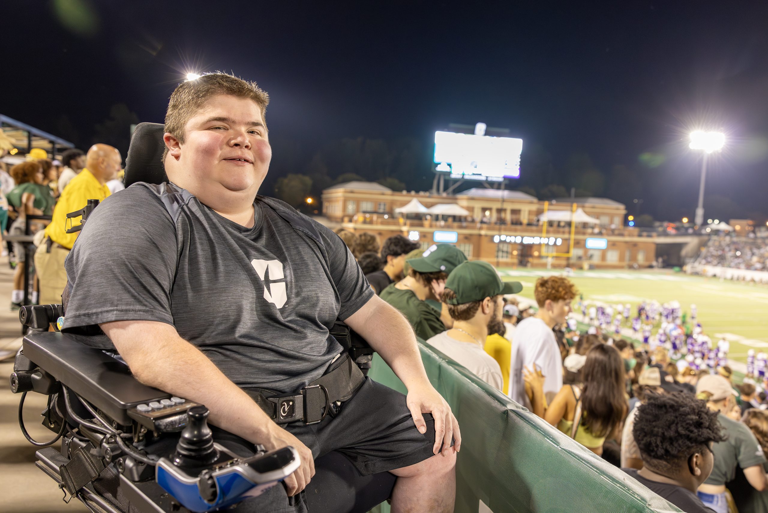 Person in a wheelchair at a sports stadium, with a crowd and field in the background at night.