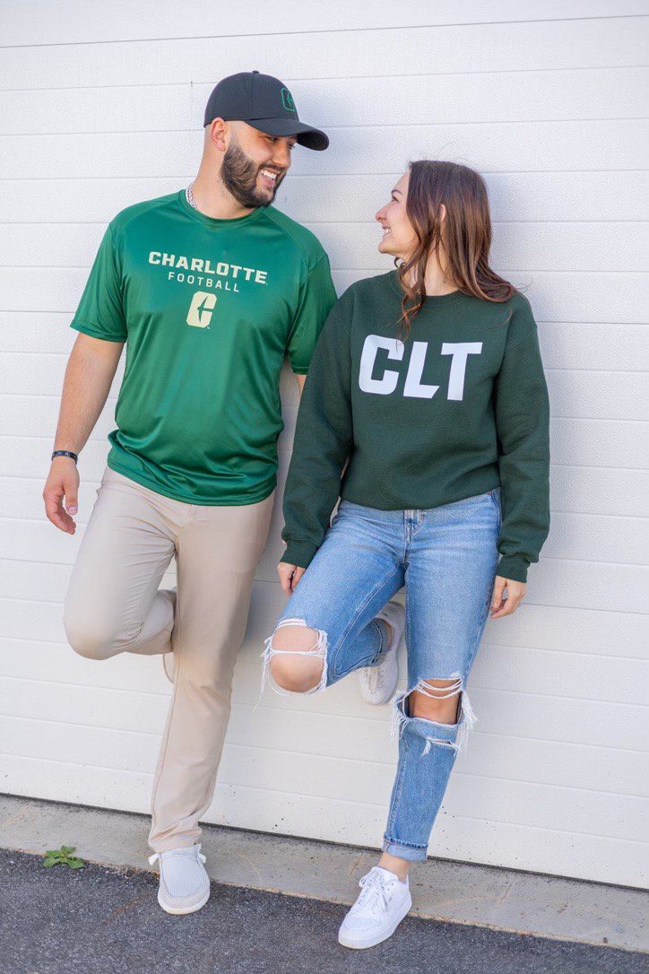 Two people in green tops and casual outfits leaning against a wall, smiling at each other.