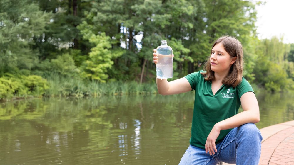 A Charlotte alumni kneels by a pond, examining a water sample in a clear bottle.