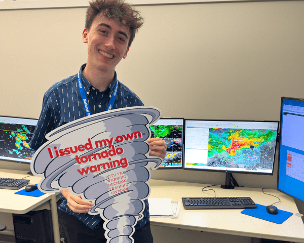 Ian Shank holding a sign in the shape of a tornado that reads "I Issued My First Tornado Warning"